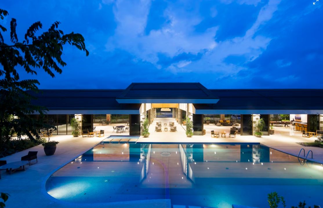 A pool area at dusk with a large pool, lounge chairs, and a building in the background.