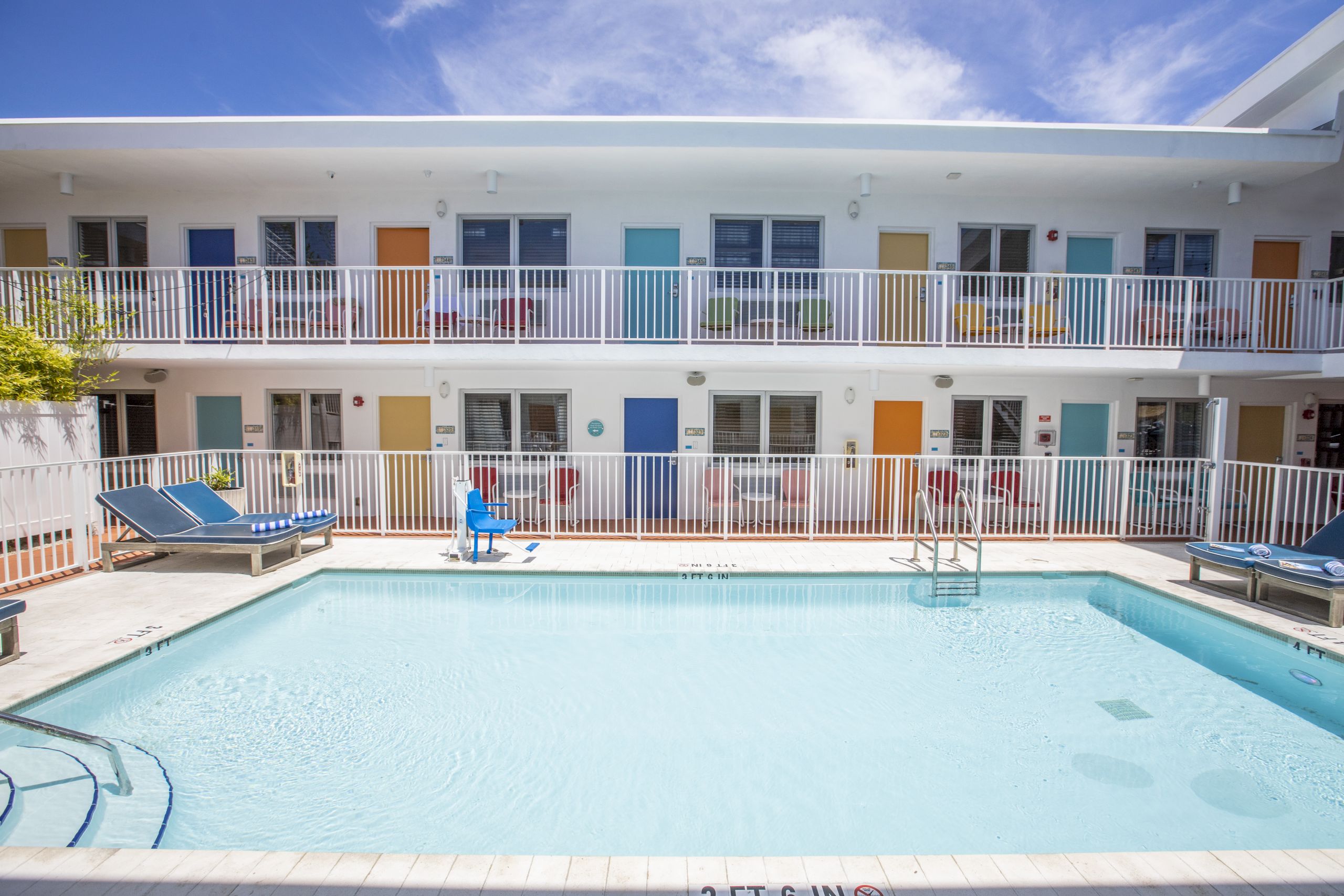 A two-story white building with colorful doors and balconies overlooking a swimming pool with lounge chairs.